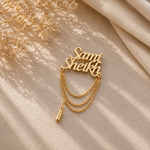 A gold-tone personalized nameplate necklace spelling 'Sammi Sheikh' in cursive, adorned with three draped chain links and a small cylindrical charm, styled on a beige textured fabric with dried flowers and soft shadows.
