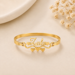 A gold-plated name bracelet with the name 'Zara' in cursive script, decorated with three small hearts and heart-shaped links, displayed on a light beige surface with dried flowers.