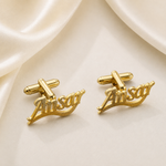 A pair of gold cufflinks shaped like stylized script names, displayed on a soft cream satin fabric background.