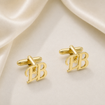 A pair of gold initial cufflinks featuring the letters 'EB' in a polished script font, displayed on a draped cream satin fabric background.
