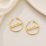 A pair of gold hoop earrings with the word 'custom' in script across the front, displayed on a soft beige fabric background.