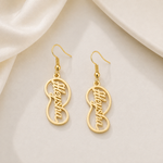 A pair of gold dangling earrings with an infinity-shaped outline and the name 'Sushant' written in cursive script inside, displayed on a soft cream satin fabric background.