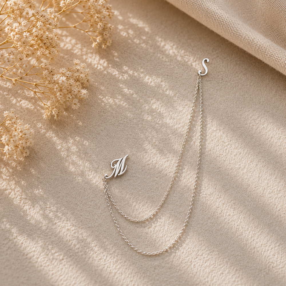A delicate silver double-chain necklace with an initial 'M' pendant and an 'S' clasp, styled on a textured beige fabric with dried flowers and soft shadows.