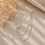 A delicate silver double-chain necklace with an initial 'M' pendant and an 'S' clasp, styled on a textured beige fabric with dried flowers and soft shadows.