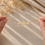 A gold necklace with Arabic calligraphy lettering is held between two hands against a beige fabric backdrop with soft shadows and dried flowers in the corner.