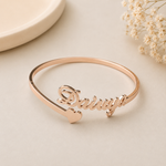A rose gold nameplate open cuff bracelet with the name 'Daiwa' in cursive and a small heart at the end, displayed on a light beige surface with dried flowers in the background.