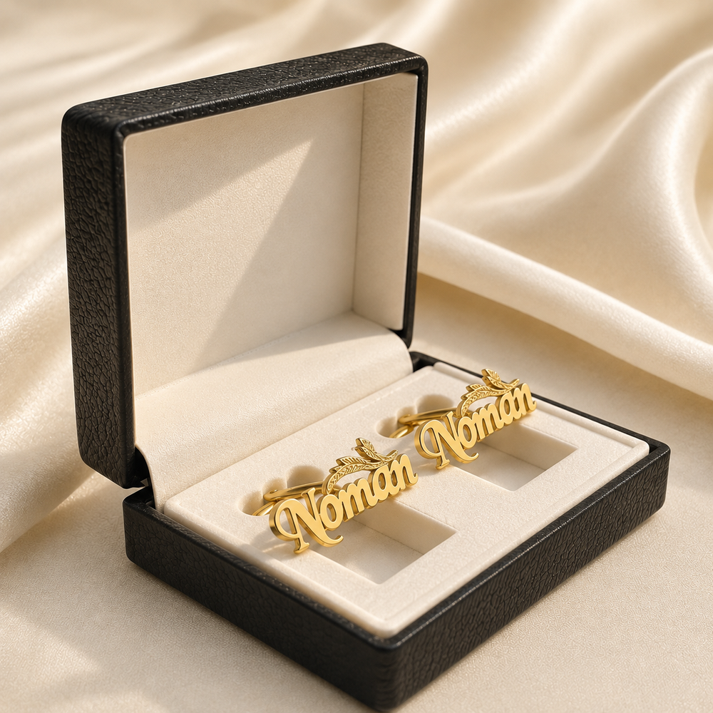 A pair of gold script cufflinks spelling 'Woman' with small heart details, presented in an open black jewelry box with a cream satin lining, set against a draped beige satin background.