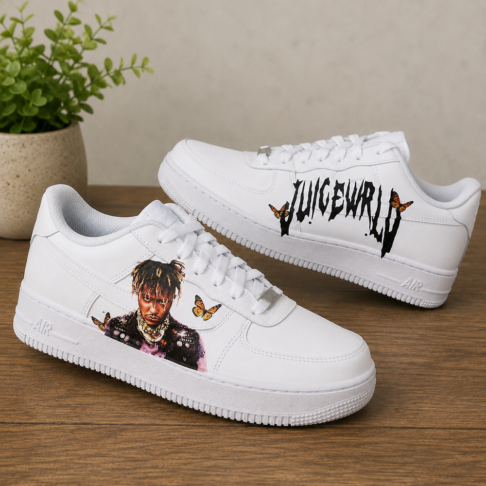 A pair of white low-top sneakers displayed on a wooden surface against a neutral beige wall. One shoe shows a printed portrait with butterflies on the side, and the other displays black graffiti-style text with butterfly accents.
