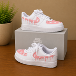 A pair of white Nike Air Force 1 Low sneakers with pink dripping graphic details, displayed on a wooden surface with one shoe resting on top of a gray Nike box and the other in front. A potted plant is visible in the background.